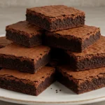 Stack of freshly baked brownies on a white plate, with a rich, fudgy texture.