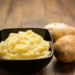 Creamy mashed potatoes in a black bowl with Yukon Gold potatoes in the background.