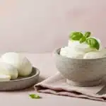 Bowl of stracciatella cheese beside mozzarella block on linen