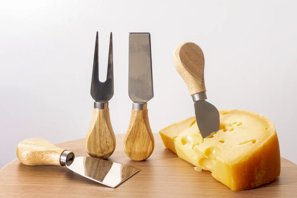 Close-up of a cheese knife with a stainless steel blade and wooden handle, placed next to assorted cheeses.
