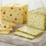 Sliced Havarti cheese with herbs and red pepper on a wooden board.