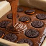 Close-up of Oreo cookies layered into glossy chocolate brownie batter in a parchment-lined pan.