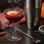 Ruby-red poisoned apple Halloween cocktail in coupe glass with dry ice fog, styled on dark wood with coral-pink linen and blue pumpkin accents.